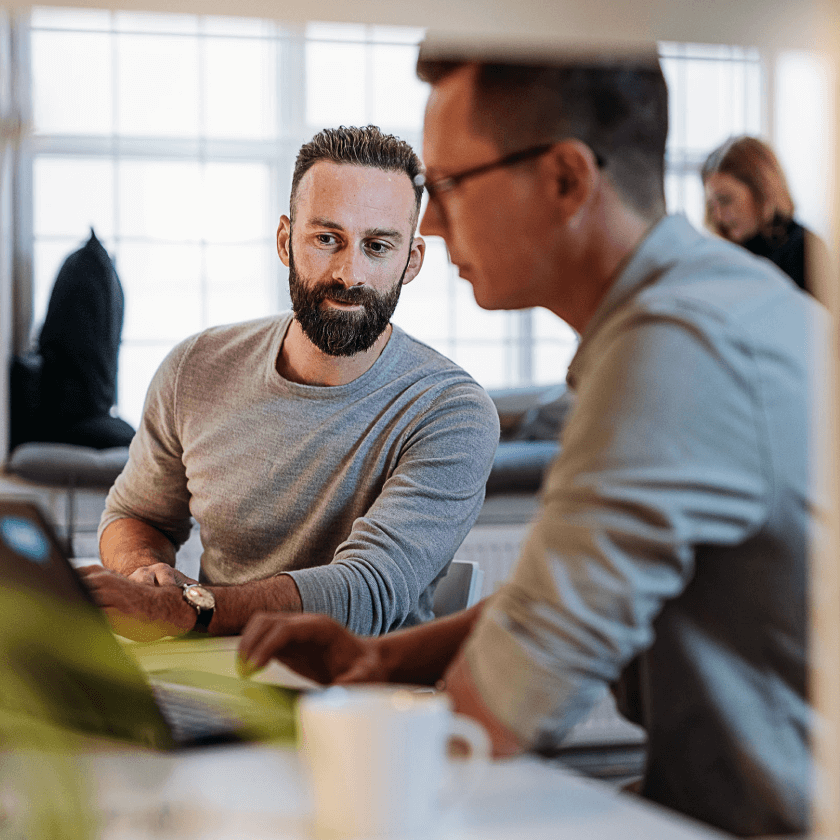 Zwei Männer diskutieren am Tisch über Docusign, während ein Laptop und eine Tasse Kaffee sichtbar sind.