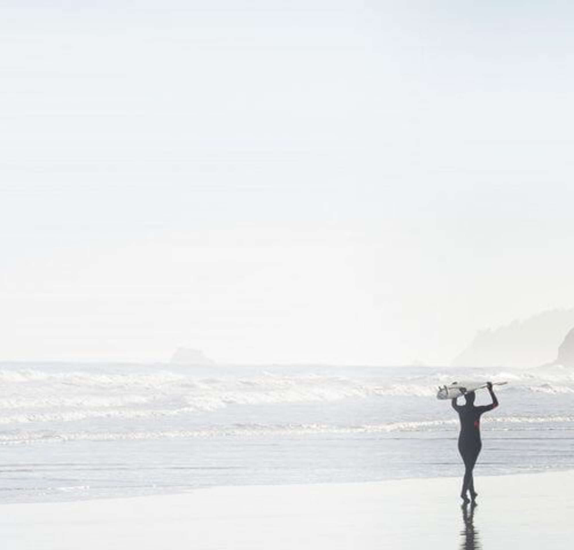 Two surfers walking together on a misty morning along a beach, carrying their surfboards above their heads.
