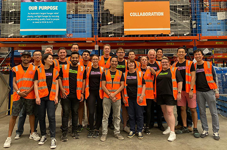 A group of 20 DocuSign employees wearing DocuSign IMPACT t-shirts and orange work vests, posing for a photo during a volunteer event in a donation warehouse 